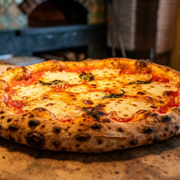 Margherita pizza with bubbly crust on a wooden board, with a pizza oven in the background.