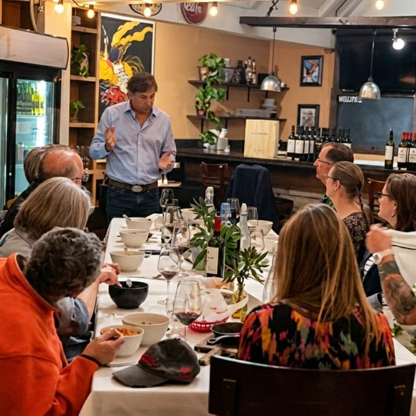 Speaker engaging a group of guests at a long dining table in a warm restaurant setting.
