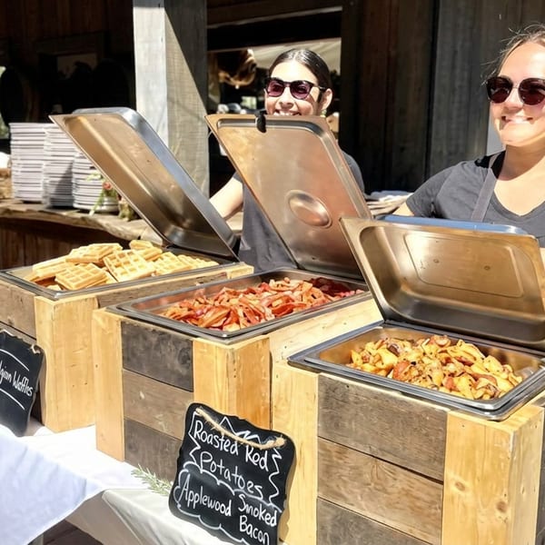 Two smiling women serve waffles, bacon, and potatoes from a rustic outdoor buffet.
