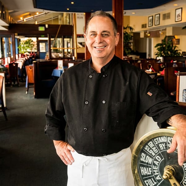 Smiling chef in black uniform leaning on a ship's telegraph in a blue and white restaurant dining room.