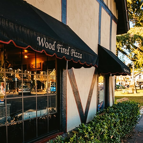 Tudor-style "Wood Fired Pizza" restaurant exterior with black awnings, reflecting a sunny street and people walking.