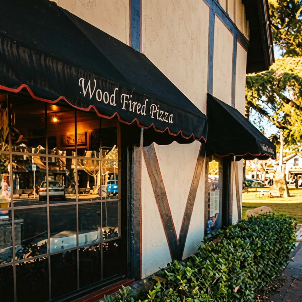 Tudor-style "Wood Fired Pizza" restaurant exterior with black awnings, reflecting a sunny street and people walking.
