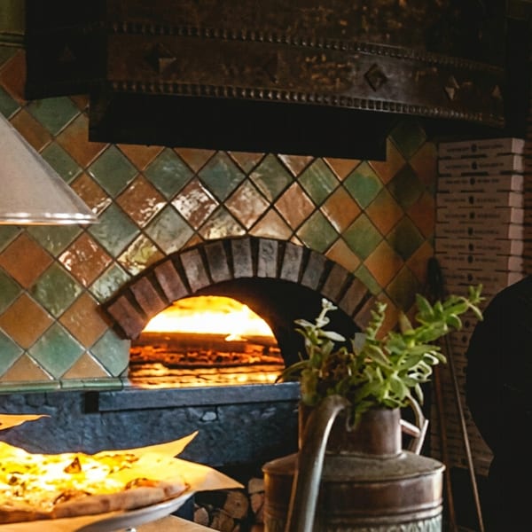 Chef near a tiled wood-fired pizza oven, with two pizzas on the counter.